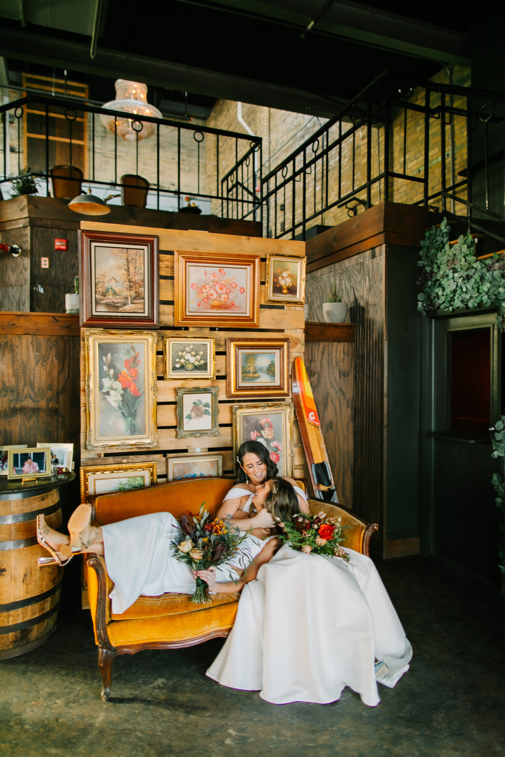 Rachel and Suzan are on a yellow couch in front of wooden pallet wall of vintage paintings and a pair of skis. Suzan lays across Rachels lap while holding her wedding bouquet and Rachel smiles down at Suzan. Their wedding is at the Cooperage in Milwaukee, Wisconsin.