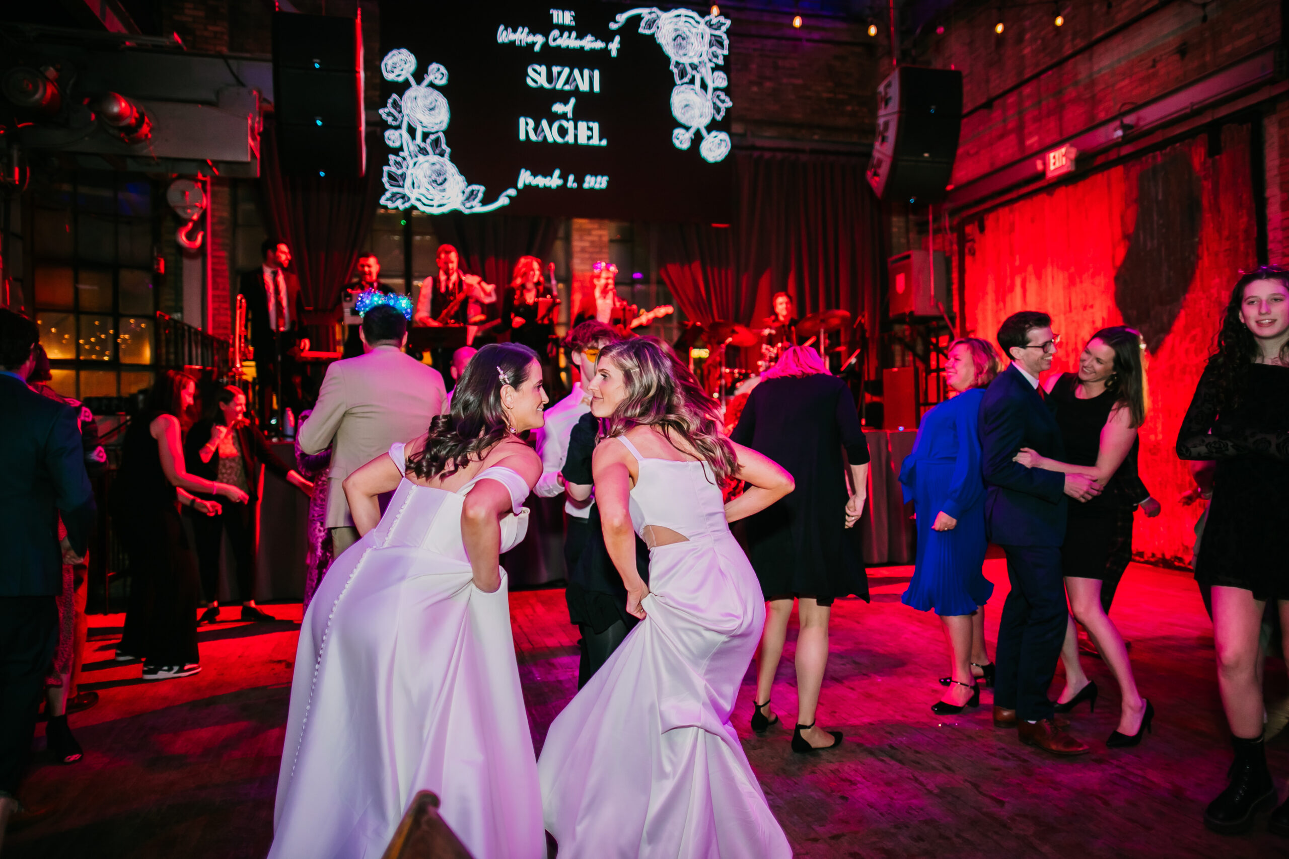 Suzan and Rachel hold their wedding dress skirts up and dance in front of the live music on stage during their wedding at The Cooperage Milwaukee.