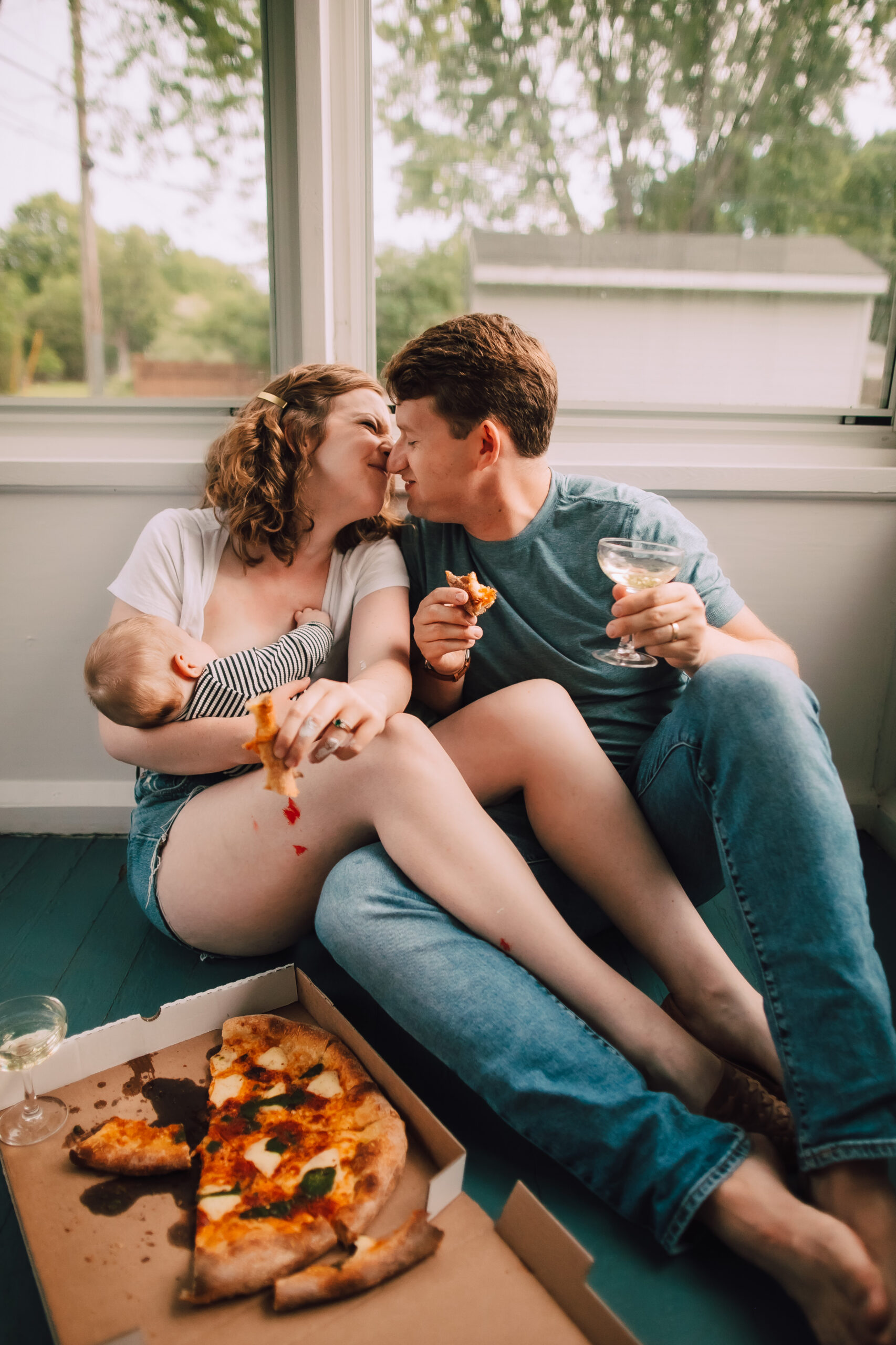 Kate and Sam lean in for a kiss while she feeds her baby and eats pizza and he eats pizza and drinks Champagne. A charming, candid moment during their new home lifestyle family photo session with O and B Photo Co in Madison, Wisconsin.